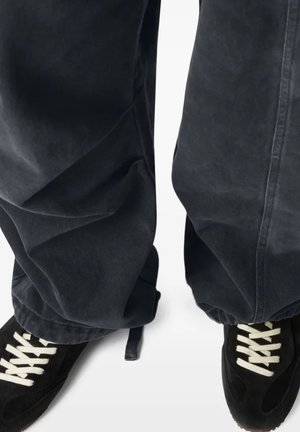 Pantaloni neri larghi che coprono le caviglie sopra sneaker nere con lacci bianchi su uno sfondo bianco.