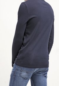 Mężczyzna w granatowej koszuli z długim rękawem i niebieskich dżinsach, lekko obrócony, z widokiem na plecy i prawą stronę, na białym tle.