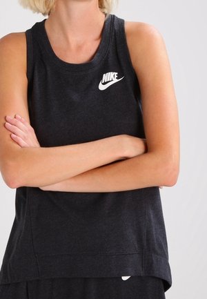 Person iført en sort ærmeløs Nike-top med armene krydset over brystet, synlig fra nakke til talje mod en ensfarvet baggrund.