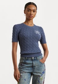 Pull à manches courtes en tricot torsadé bleu marine avec un ourlet côtelé et un détail de logo brodé sur le côté gauche. Associé à un jeans éclaboussé de peinture.