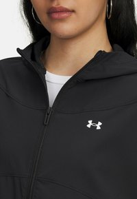 Melnā kapucēta jakā, izgatavota no mīksta auduma, ar pilnu rāvējslēdzēju un baltu Under Armour logotipu kreisajā pusē. Tuvplāns uz apkakles.