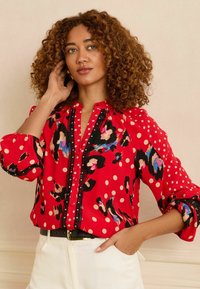 Sarkans blūze ar melnu un krāsainu dzīvnieku rakstu, klasiskām punktiņām un mežģīņu apdari. Ar garām pūkainām piedurknēm un pogu aizdari priekšpusē.