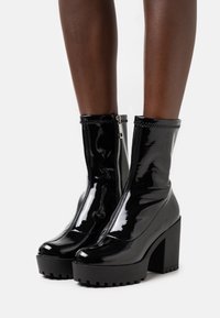 Bottes ankle en cuir verni noir brillant avec des semelles compensées épaisses et des zips latéraux portées sur des jambes à la peau foncée sur fond blanc.