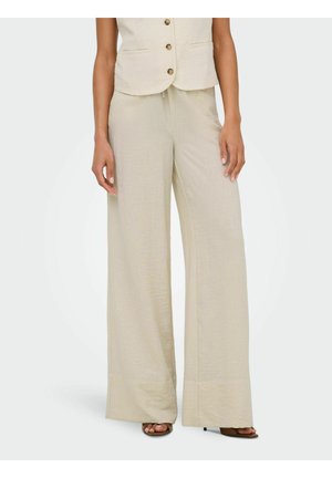 Femme portant un pantalon en lin beige taille haute à jambes larges et un gilet boutonné assorti, debout, les mains détendues le long du corps.