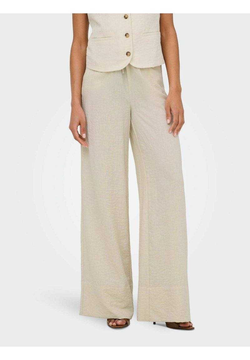 Femme portant un pantalon en lin beige taille haute à jambes larges et un gilet boutonné assorti, debout, les mains détendues le long du corps.