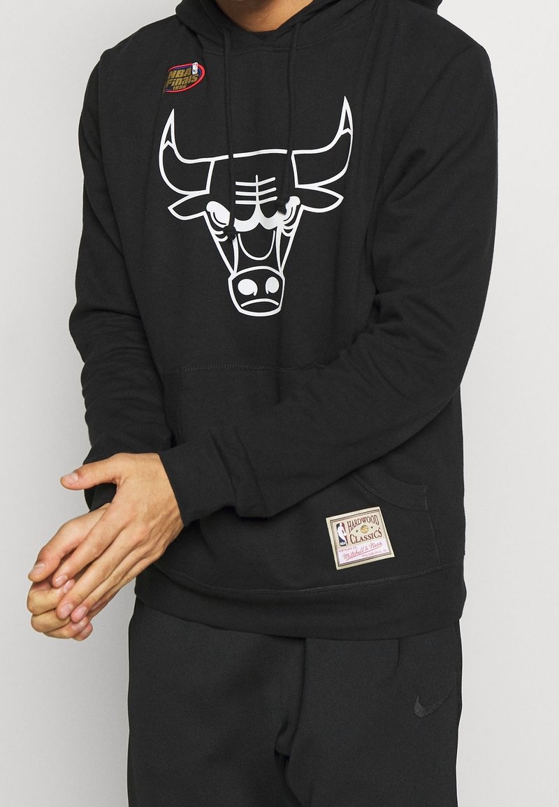 Person som bär en svart huvtröja med en vit Chicago Bulls-logotyp och en NBA Finals 1996-lapp, i kombination med svarta Nike-byxor.