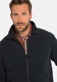 Giacca in pile blu navy con collo alto, zip frontale e due tasche laterali, indossata sopra una camicia a quadri rossa e bianca.