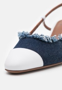 Chaussure en tissu denim bleu avec des détails à bords effilochés, capuchon de orteil en cuir blanc et semelle intérieure en cuir brun. Sangle ajustable au talon pour un maintien sécurisé.