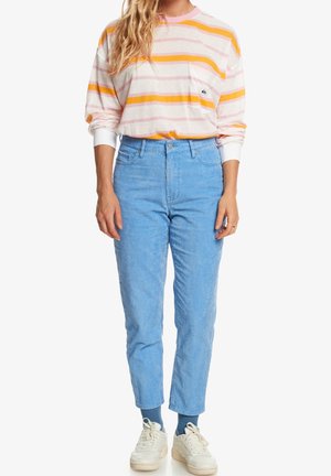Vrouw draagt lichtblauwe corduroy broek, witte sneakers, blauwe sokken en een hemd met lange mouwen met oranje en roze strepen en een borstzak.