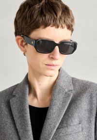 Black rectangular sunglasses with dark lenses, matte finish, and gold logo on the arm; worn with a grey wool blazer and black top.