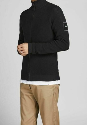 Homme portant un pull zippé noir avec un patch logo sur la manche, un pantalon beige et une chemise blanche qui dépasse en dessous.