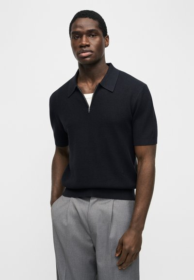 Νεαρός άνδρας που φοράει μαύρο μάλλινο πουλόβερ με φερμουάρ και κοντά μανίκια πάνω από λευκό φανελάκι και ανοιχτόχρωμο γκρι πλεκτό παντελόνι, στεκόμενος με τα χέρια στις τσέπες