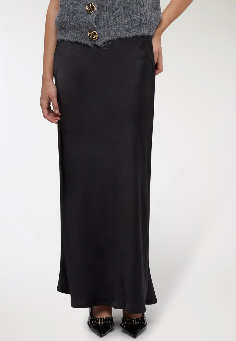 Gonna lunga in satin nero, texture liscia, design fluente. Abbinata a un top grigio peloso e décolleté nere con punta appuntita e dettagli borchiati.