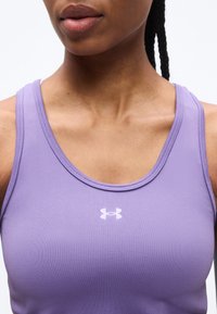 Γυναίκα με αμάνικο αθλητικό τοπ σε λιλά χρώμα με το λογότυπο Under Armour στο κέντρο του στήθους, εμφανίζει το λαιμό και μέρος του προσώπου της και πλεγμένα μαλλιά.