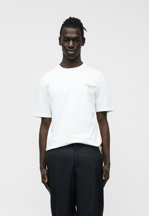 Jeune homme à la peau foncée et aux cheveux tressés, portant un t-shirt blanc à manches courtes et un pantalon noir, sur un fond uni clair.