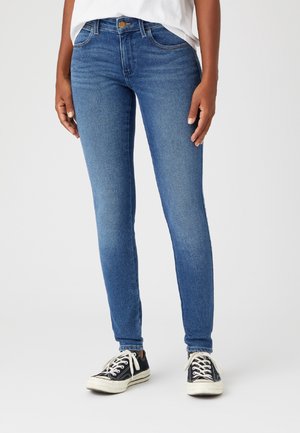 Jeans skinny de mezclilla azul con un diseño clásico de cinco bolsillos y un ligero desgastado. Combinados con zapatillas negras con cordones blancos.