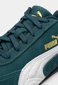Baskets en daim vert avec des accents en cuir blanc, lacets plats et logo PUMA doré. Présentent un design texturé sur la semelle et la tige.
