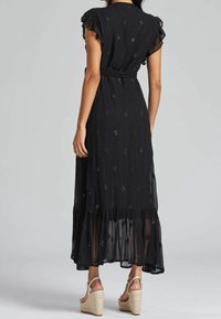 Schwarzes Maxi-Kleid mit rüschierten Ärmeln, offener Rückseite und dekorativen Diamantmustern. Hat einen geschichteten Saum und wird mit Wedge-Sandalen kombiniert.