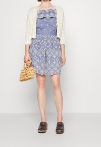 Hellblaues Kleid mit weißer Stickerei und gewellten Kanten, kombiniert mit einer beigen Strickjacke und braunen Clogs. Accessoire: geflochtene Holzhandtasche.