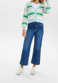 Kvinna i randig kardigan i grönt, vitt och blått, högmidjade vida blå jeans och vita sneakers, stående mot en vit bakgrund.