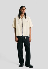Persona con lunghi capelli scuri indossa una camicia beige a maniche corte con bottoni, pantaloni neri a gamba larga con portachiavi e mocassini neri su uno sfondo grigio.