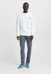 Felpa bianca a girocollo con piccolo logo blu, abbinata a jeans grigi e sneakers blu, con strisce bianche sui lati.