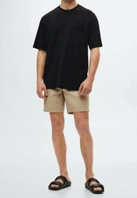 Person iført sort kortærmet t-shirt, beige shorts og sorte dobbelte remsandaler står foran en ensfarvet baggrund.