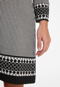 Schwarz-weißes gemustertes Kleid mit Hahnentrittmuster, das am Saum mit einem geometrischen Rand und einem Manschettendetail mit sechseckigem Design versehen ist.