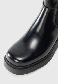 Bottillon en cuir noir avec une finition brillante, bout arrondi, semelle épaisse noire et panneaux latéraux élastiques pour un enfilage facile.