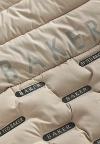 Beiger Puffer-Jacke aus einem gesteppten Stoff. Großes silbernes "BAKER"-Logo und kleine schwarze Etiketten mit den Aufdrucken "BAKER" und "BY TED BAKER".