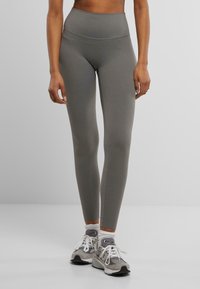 Leggings taille haute en matériau gris uni, présentant une texture lisse et un design ajusté, portés avec des baskets grises et des chaussettes blanches à mi-mollet.