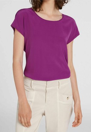 Fioletowa bluzka z krótkim rękawem o okrągłym dekolcie i gładkiej fakturze, zestawiona z jasnobeżowymi spodniami z kieszeniami z przodu i ozdobnymi elementami.