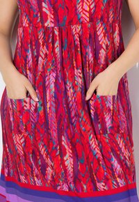 Červené šaty se živým peřím vzorem, s skládacím živůtkem a dvěma bočními kapsami. Lem šatů má vrstvené barevné pruhy v purpurové a červené barvě.
