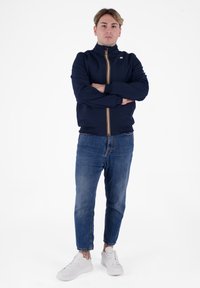Giacca navy con colletto alto, chiusura frontale con zip e dettagli a strisce arancioni, abbinata a jeans blu e sneakers bianche. Tessuto dalla consistenza liscia.