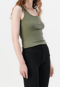 Olivgrünes, geripptes Tanktop mit figurbetontem Design, das einen runden Ausschnitt und drei Knopfdetails aufweist, kombiniert mit hochgeschnittenen schwarzen Jeans.