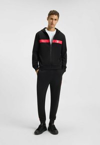Felpa nera con zip e striscia rossa sul petto, abbinata a pantaloni jogger coordinati. Tessuto morbido, vestibilità comoda e sneakers nere.