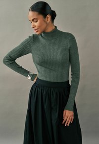 Rippstrick grüner Rollkragenpullover mit langen Ärmeln, kombiniert mit einem schwingenden schwarzen Rock. Hat eine glatte Textur und eine figurbetonte Silhouette.