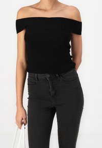 Top negro de canalé sin mangas combinado con jeans ajustados oscuros; modelo sosteniendo un bolso blanco, mostrando un look elegante y minimalista.