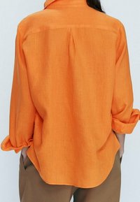 Orangenes Leinenhemd mit Kragen, hochgekrempelten Ärmeln und einer zentralen Falte am Rücken. Es hat eine lässige Passform und eine glatte Textur.