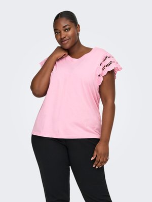 Femme souriante portant un haut rose avec des manches courtes en dentelle et un pantalon noir, posant avec une main près du cou sur un fond uni.