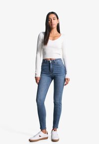 Haut court à manches longues côtelé blanc associé à un jean skinny bleu taille haute. Le mannequin porte des baskets blanches avec des accents marron. Fond simple.