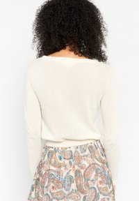 Kremowy sweter z długimi rękawami, miękką teksturą i szerokim dekoltem, zestawiony z wzorzystą spódnicą z kwiatowymi i paisley'owymi wzorami.
