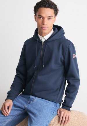 Felpa con zip di colore blu navy in tessuto morbido, con zip frontale, cordini e patch con logo sulla manica sinistra. Abbinata a jeans di colore azzurro.