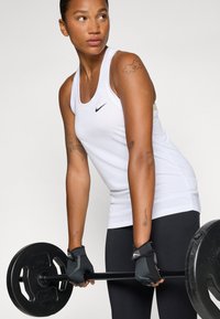 Biały sportowy top na ramiączkach z czarnym logo Nike, w zestawie z czarnymi legginsami i rękawicami do podnoszenia ciężarów, trzymający czarny talerz ciężarowy.