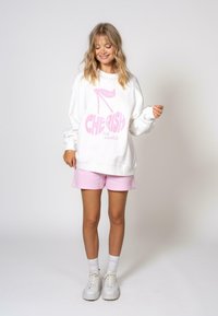 Un sweat-shirt blanc avec un texte graphique rose audacieux qui dit "CHÉRIR le moment" et un design de talon haut. Assorti à un short rose clair.