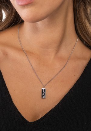 Nahaufnahme einer Person mit einer silbernen rechteckigen Anhängerkette, auf der der Schriftzug "the good" und ein kleines Herzsymbol eingraviert sind.