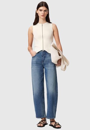 Femme portant un haut blanc sans manches à fermeture éclair sur le devant, un jean large taille haute bleu, des sandales noires, tenant un grand sac fourre-tout crème.