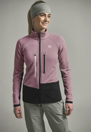 Lächelnde Frau, die eine rosa-schwarze Jacke mit Reißverschluss, graue Hose und graues Stirnband trägt, steht vor einem einfarbigen Hintergrund.