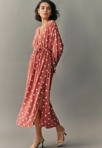 Rochie lungă, de culoare roz aprins, cu buline albe, având decolteu în V, mâneci lungi, talie strânsă cu șnur și o fanta pe lateral.