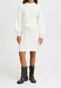 Robe tricotée blanche avec de longues manches bouffantes et une ceinture à la taille, présentant une fente sur le côté et une texture côtelée. Assortie à des bottes noires jusqu'aux genoux.